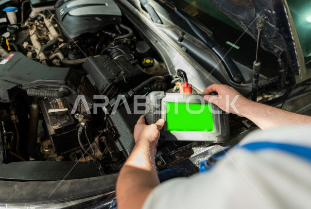 رجل عربي سعودي  داخل ورشة السيارات ، يستخدم جهاز التابلت بخلفية خضراء كروما لاختيار خدمة التصليح ، مهندس ميكانيكي يقوم باصلاح سيارته ، استخدام ادوات للصيانة ، اصلاح يدوي ، خدمات اصلاح و صيانة السيارات ، مهندس ميكانيكي ، ورشة صيانة 