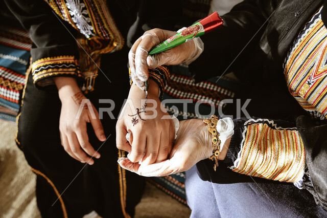 عادات وتقاليد في المناسبات والأفراح السعيدة، صورة مقربة ليد إمرأة عربية خليجية سعودية ترتدي الزي التقليدي و تقوم برسم و نقش الحنة على يد فتاة، مهن تقليدية شعبية نسائية، زخارف وفنون يدويه من مواد طبيعيه، التجمل والاهتمام بالمظهر الخارجي على الطريقة الشعبية