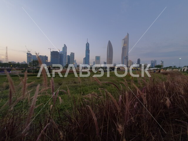 الأبراج المطلة على حديقة الشهيد في منطقة الكويت الثقافية الوطنية الجديدة، الاهتمام بإضاءة وإنارة المناطق والأماكن العائلية للتنزه والمتعة، ممرات المشي والتجول، منظر المروج الخضراء والأشجار والبيئة الطبيعية وقت الغروب، المنتزهات والحدائق العامة في دولة الكويت