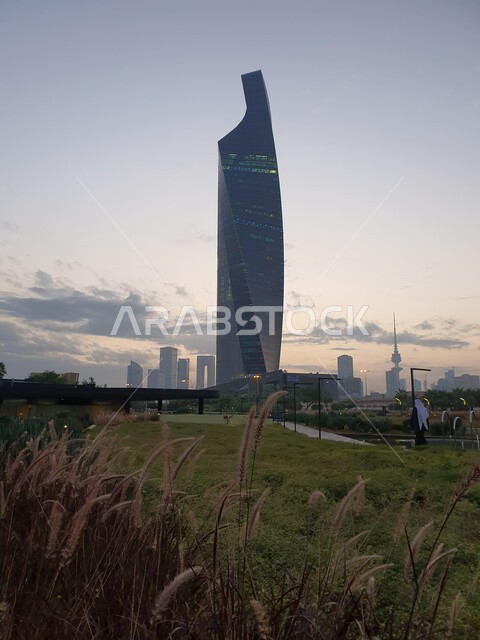  برج التجارية المطل على حديقة الشهيد في منطقة الكويت الثقافية الوطنية الجديدة، المناطق والأماكن العائلية للتنزه والمتعة، ممرات المشي والتجول، منظر المروج الخضراء والأشجار والبيئة الطبيعية وقت الغروب، المنتزهات والحدائق العامة في دولة الكويت