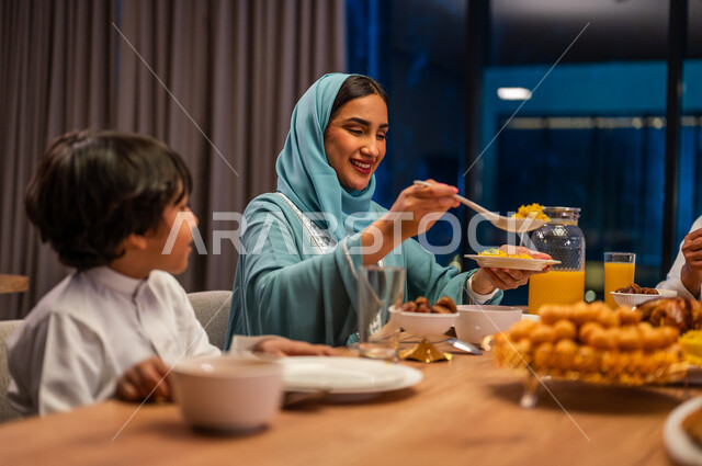 مائدة طعام بأصناف متنوعة، صورة مقربة لإمرأة عربية خليجية سعودية تقوم بسكب الأطعمة تجلس بجانب ابنها، أجواء عائلية سعيدة في شهر رمضان المبارك، فوانيس معدنية مضيئة وديكورات وزينة المناسبات والأعياد الاسلامية 