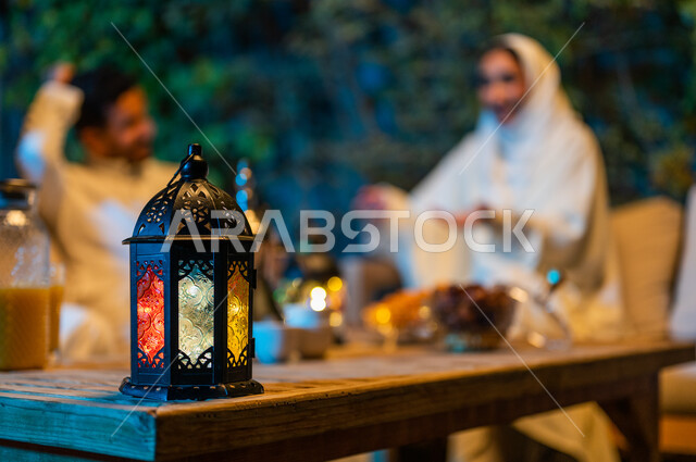 ديكورات وزينة المناسبات والأعياد الاسلامية، تبادل الأحاديث والتسامر بين الأفراد، أسرة عربية خليجية سعودية تقضي أوقات ممتعة في جلسة خارجية في احدى الشاليهات، أجواء عائلية سعيدة في شهر رمضان المبارك، فانوس معدني مضيء على طاولة خشبية 
