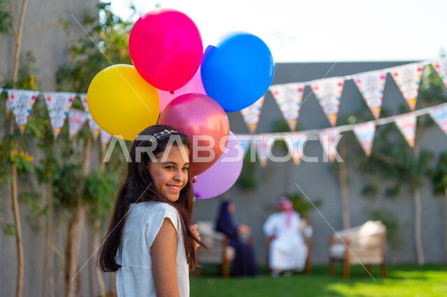 Preparing to celebrate happy Islamic occasions, the joy and happiness of children to welcome Eid, holiday customs in Saudi Gulf society, a close-up photo from the side of a Saudi Gulf Arab girl wearing a white dress, holding colorful balloons in her hand with gestures of happiness