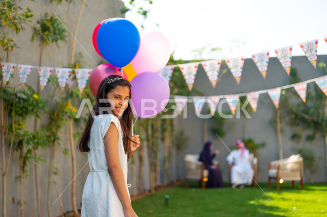 ديكورات وزينة في إحدى الشاليهات والمنتجعات وقت الأعياد في المجتمع العربي السعودي, فتاة عربية سعودية خليجية ترتدي فستان أبيض تحمل بيدها بالونات ملونة بايماءات السرور, التجهيز للاحتفال بالمناسبات الإسلامية السعيدة، بهجة وفرحة الأطفال لاستقبال العيد