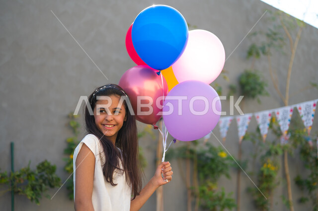 The joy and happiness of children to welcome Eid, holiday customs in Saudi Gulf society, a close-up photo of a Saudi Gulf Arab girl wearing a white dress, holding colorful balloons in her hand with gestures of happiness, preparing to celebrate happy Islamic occasions