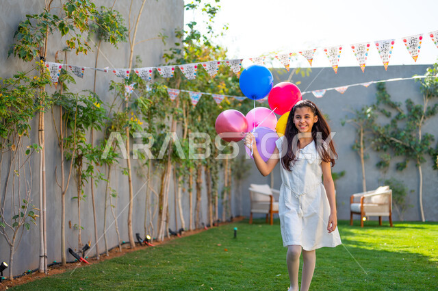 ديكورات وزينة في إحدى الشاليهات والمنتجعات وقت الأعياد في السعودي, التجهيز للاحتفال بالمناسبات الإسلامية السعيدة، بهجة وفرحة الأطفال لاستقبال العيد، فتاة عربية سعودية خليجية ترتدي فستان أبيض تحمل بيدها بالونات ملونة بايماءات السرور, صورة الجسم بالطول الكامل