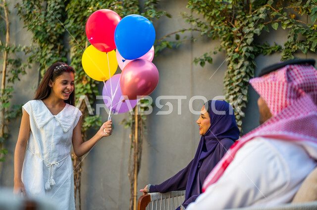 قضاء أوقات عائلية ممتعه, المناسبات والأعياد الدينية, عائلة سعودية تستمتع بالهواء الطلق, سرور وفرحة الأطفال بقدوم العيد, فتاة عربية خليجية سعودية ترتدي الفستان الأبيض تمسك بيدها مجموعة من البلالين الملونة بفرح وبشاشة, الأجواء الأسرية السعيدة