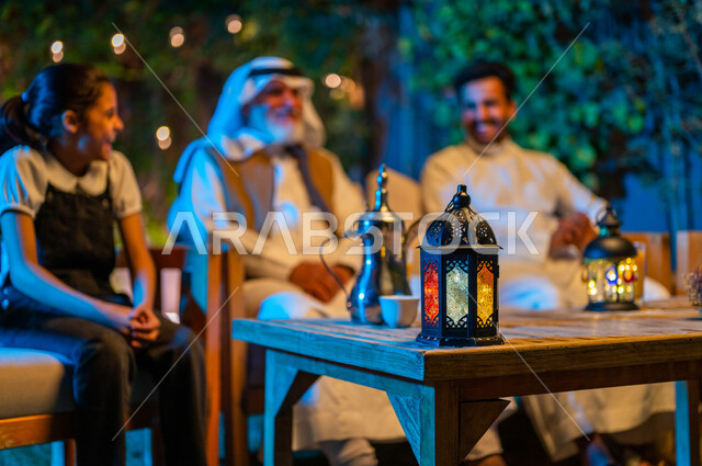ديكورات وزينة المناسبات والأعياد الاسلامية، تبادل الأحاديث والتسامر بين الأفراد، أسرة عربية خليجية سعودية تقضي أوقات ممتعة في جلسة خارجية في احدى الشاليهات، أجواء عائلية سعيدة في شهر رمضان المبارك، فانوس معدني مضيء على طاولة خشبية