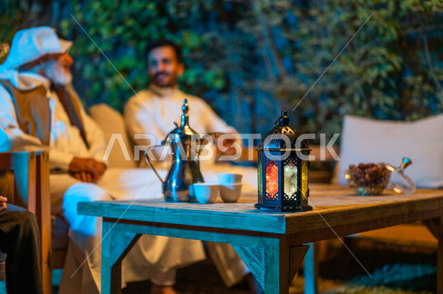 ديكورات وزينة المناسبات والأعياد الاسلامية، تبادل الأحاديث والتسامر بين الأفراد، أسرة عربية خليجية سعودية تقضي أوقات ممتعة في جلسة خارجية في احدى الشاليهات، أجواء عائلية سعيدة في شهر رمضان المبارك، فانوس معدني مضيء على طاولة خشبية 
