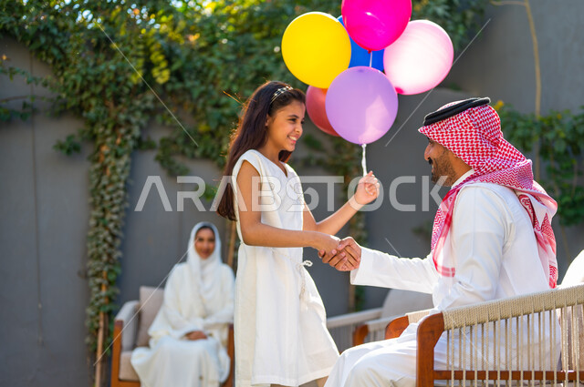 ايماءات القاء التحية والسلام, مفهوم الترابط الأسري, الاحتفال بالمناسبات والأعياد في الهواء الطلق, أجواء العيد في المجتمع السعودي, تعابير الاحترام وتقدير الكبار, صورة من الجانب لفتاة عربية خليجية سعودية ترتدي الفستان الأبيض تمسك بلالين تقوم بالسلام على والدها