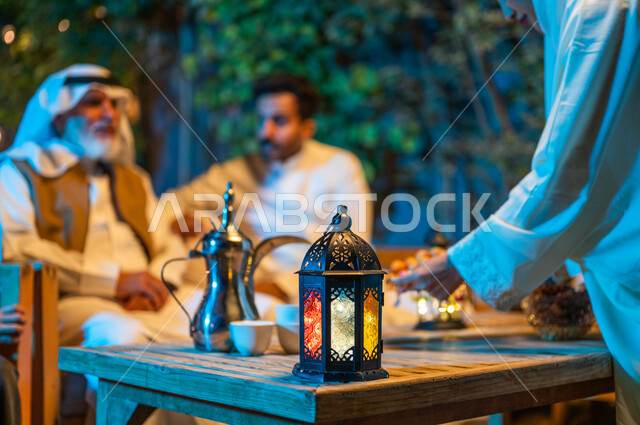أجواء عائلية سعيدة في شهر رمضان المبارك، تبادل الأحاديث والتسامر بين الأفراد، أسرة عربية خليجية سعودية تقضي أوقات ممتعة في جلسة خارجية في احدى الشاليهات، صورة مقربة ليد امرأة تقوم بوضع حلوى اللقيمات على طاولة خشبية، فوانيس معدنية مضيئة