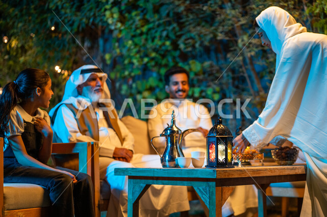 أجواء عائلية سعيدة في شهر رمضان المبارك، تبادل الأحاديث والتسامر بين الأفراد، أسرة عربية خليجية سعودية تقضي أوقات ممتعة في جلسة خارجية في احدى الشاليهات، سيدة  تقوم بوضع حلوى اللقيمات على طاولة خشبية، فوانيس معدنية مضيئة