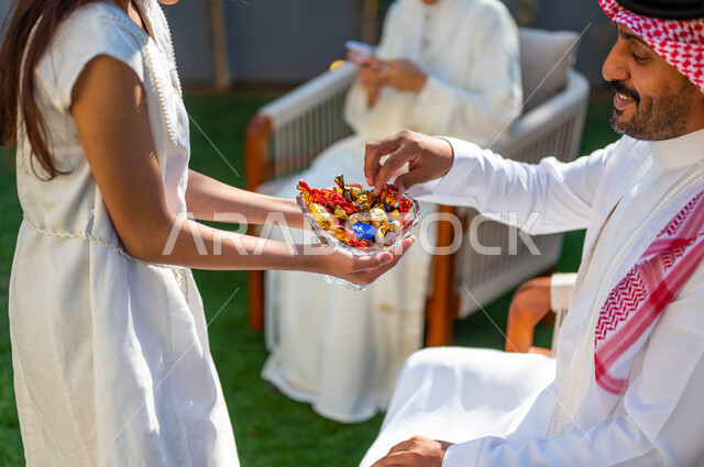 الاحتفال بالهواء الطلق في العيد المبارك, فرحة و بهجة الأطفال في العيد السعيد، صورة مقربة لفتاة عربية خليجية سعودية ترتدي فستان أبيض تقوم بتقديم قطع السكاكر, عادات العيد في المجتمع السعودي الخليجي، أعياد ومناسبات إسلامية سعيدة