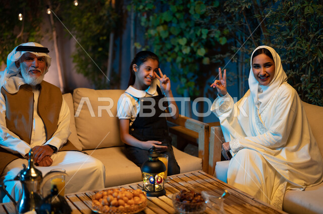 A Saudi Gulf Arab family looking at the camera spending enjoyable times in an outdoor session in one of the chalets, decorations and decorations for Islamic occasions and holidays, exchanging conversations and socializing between individuals, a happy family atmosphere in the holy month of Ramadan, a mother and daughter raising their hands with happy gestures