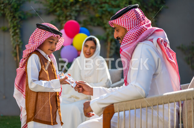العادات والتقاليد العربية في العيد، إدخال الفرحة لقلوب الأطفال،  قضاء وقت ممتع في أيام العيد مع الأسرة، ارتداء الأزياء التقليدية في المناسبات والأعياد الإسلاميه، صورة منالجانب لولد عربي سعودي خليجي يرتدي الزي التقليدي يقوم بأخذ العيدية من والده
