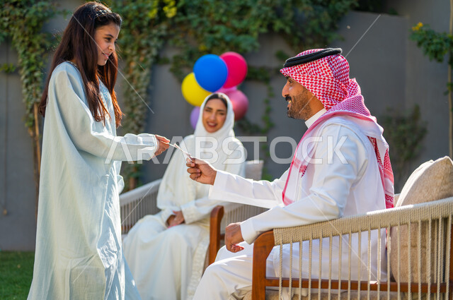 قضاء وقت ممتع مع العائلة في أيام العيد، أجواء وطقوس العيد في المجتمع العربي السعودي, إدخال الفرحة لقلوب الأطفال،  صورة من الجانب لفتاة عربية سعودية خليجية ترتدي الفستان الأبيض تقوم بأخذ العيدية من والدها,ارتداء الأزياء التقليدية في المناسبات والأعياد الإسلاميه