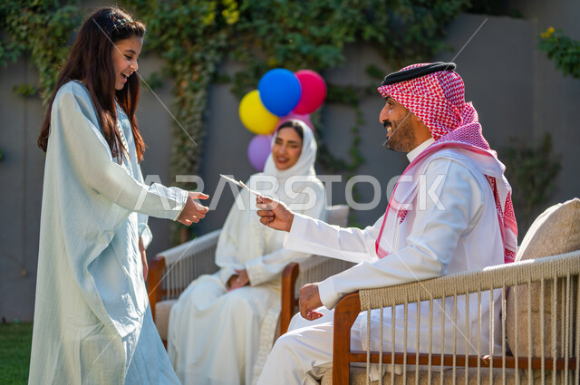 The atmosphere and rituals of Eid in Saudi Arabian society, bringing joy to the hearts of children, spending quality time with the family on Eid days, wearing traditional costumes on Islamic occasions and holidays, a picture from the side of a Saudi Arabian Gulf girl wearing a white dress, taking Eid from her father.