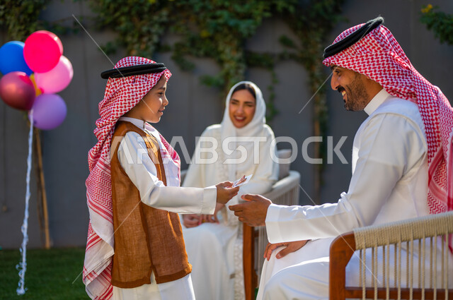 إدخال الفرحة لقلوب الأطفال، طقوس عادات وتقاليد عربية في العيد، قضاء وقت ممتع في أيام العيد مع الأسرة، ارتداء الأزياء التقليدية في المناسبات والأعياد الإسلاميه، صورة منالجانب لولد عربي سعودي خليجي يرتدي الزي التقليدي يقوم بأخذ العيدية من والده