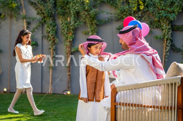 إيماءات وتعابير السعادة والسرور، ارتداء الأزياء التقليدية في المناسبات والأعياد الإسلاميه، صورة من الجانب لرجل عربي سعودي خليجي يرتدي الزي التقليدي يقوم بتعديل شماغ ابنه, مفهوم الأناقة والاهتمام بالمظهر الخارجي، قضاء وقت ممتع في أيام العيد في السعوديه مع العائلة