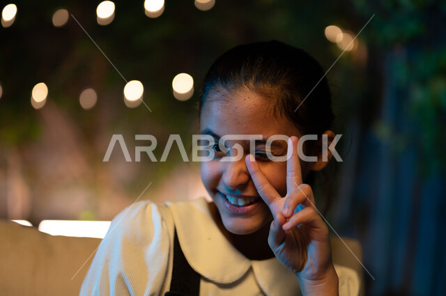 الأعياد والمناسبات الدينية الاسلامية، صورة مقربة لفتاة عربية خليجية سعودية مبتسمة تنظر إلى الكاميرا ترفع يدها بإيماءات الفرح والسرور، الاستمتاع بقضاء أوقات متميزة في جلسة خارجية في احدى الشاليهات