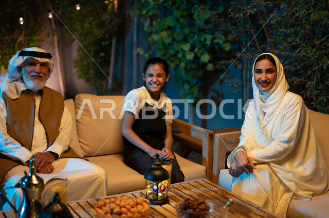 النظر إلى الكاميرا بإيماءات الفرح والسرور، ديكورات وزينة المناسبات والأعياد الاسلامية، أسرة عربية خليجية سعودية تقضي أوقات ممتعة في جلسة خارجية في احدى الشاليهات، أجواء عائلية سعيدة في شهر رمضان المبارك، فانوس معدني مضيء على طاولة خشبية