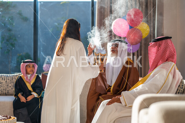 التطييب والتعطر بالعود والبخور في الأعياد والمناسبات، أسرة عربيه خليجية سعودية تجتمع في غرفة الجلوس احتفالا بالعيد، ارتداء الملابس التقليدية، ايماءات البهجة والسرور
