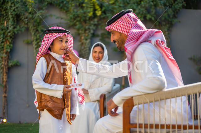 طقوس وعادات المجتمع السعودي في توزيع العيديات، صورة من الجانب لرجل عربي سعودي خليجي يقوم بملاطفة ابنه بايماءات السعادة والسرور، إدخال الفرحة والبهجة لقلوب الأولاد، المناسبات والأعياد الإسلامية، قضاء وقت ممتع مع العائلة