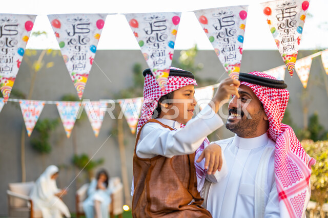 مفهوم الأناقة والاهتمام بالمظهر الخارجي للاحتفال في العيد، ارتداء الزي الشعبي في المناسبات والأعياد الإسلاميه في السعوديه، قضاء وقت ممتع مع العائلة، صورة مقربة لرجل عربي سعودي خليجي يرتدي الشماغ والثوب التقليدي يحمل ابنه يقوم بمسك الزينة بتعابير الفرح والسرور