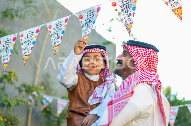 ارتداء الزي الشعبي في المناسبات والأعياد الإسلاميه في السعوديه، قضاء وقت ممتع مع العائلة، تعابير الفرح والسرور, مفهوم الأناقة والاهتمام بالمظهر الخارجي للاحتفال في العيد، صورة مقربة لرجل عربي سعودي خليجي يرتدي الشماغ والثوب التقليدي يحمل ابنه يقوم بمسك الزينة
