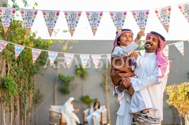 التجمعات الأسريه في المناسبات للاحتفال بالاعياد الاسلامية, أجواء العيد في المجتمع العربي السعودي، رجل عربي خليجي سعودي مبتسم يرتدي الشماغ والثوب التقليدي يحمل ابنه ويرفعه عاليا، تعابير وايماءات الفرح والسرور، زينة وديكورات الأعياد في الشاليهات