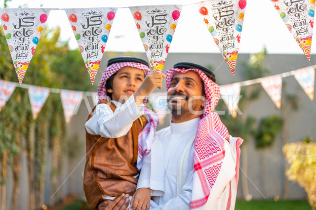 قضاء وقت ممتع مع العائلة، تعابير الفرح والسرور، ارتداء الزي الشعبي في المناسبات والأعياد الإسلاميه في السعوديه، صورة مقربة لرجل عربي سعودي خليجي يرتدي الشماغ والثوب التقليدي يحمل ابنه يقوم بمسك الزينة, مفهوم الأناقة والاهتمام بالمظهر الخارجي للاحتفال في العيد