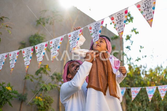 Spending quality time with the family, expressions of joy and happiness, wearing popular costumes on Islamic occasions and holidays in Saudi Arabia, a close-up picture of a Saudi Gulf Arab man wearing a shemagh and traditional thobe carrying his son holding decorations, the concept of elegance and attention to the external appearance of celebrating the holiday.
