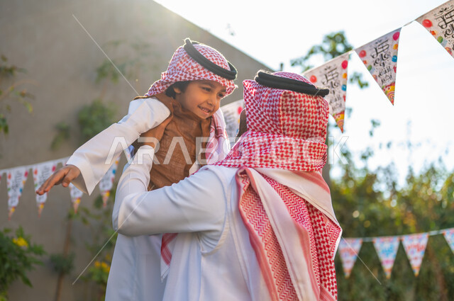 زينة وديكورات الأعياد في الشاليهات, التجمعات الأسريه في المناسبات للاحتفال بالاعياد الاسلامية, أجواء العيد في المجتمع العربي السعودي، صورة من الجانب لرجل عربي خليجي سعودي مبتسم يرتدي الشماغ والثوب التقليدي يحمل ابنه ورفعه عاليا، تعابير وايماءات الفرح والسرور
