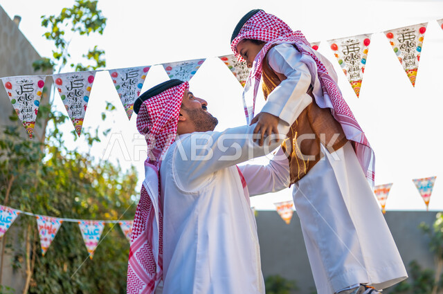 أجواء العيد في المجتمع العربي السعودي، التجمعات الأسريه في المناسبات للاحتفال بالاعياد الاسلاميةصورة من الجانب لرجل عربي خليجي سعودي مبتسم يرتدي الشماغ والثوب التقليدي يحمل ابنه ورفعه عاليا، تعابير وايماءات الفرح والسرور، زينة وديكورات الأعياد في الشاليهات