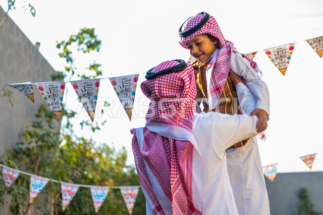 زينة وديكورات الأعياد في الشاليهات, أجواء العيد في المجتمع العربي السعودي، صورة من الخلف لرجل عربي خليجي سعودي يرتدي الشماغ والثوب التقليدي يحمل ابنه ويرفعه عاليا، تعابير وايماءات الفرح والسرور, التجمعات الأسريه في المناسبات للاحتفال بالاعياد الاسلامية 