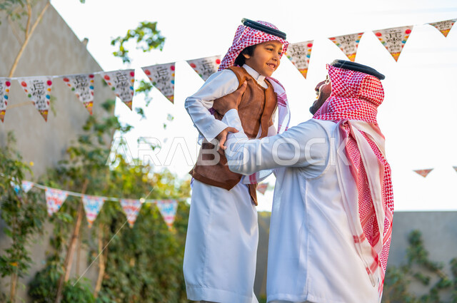 التجمعات الأسريه في المناسبات للاحتفال بالاعياد الاسلامية, أجواء العيد في المجتمع العربي السعودي، صورة من الجانب لرجل عربي خليجي سعودي مبتسم يرتدي الشماغ والثوب التقليدي يحمل ابنه ورفعه عاليا، تعابير وايماءات الفرح والسرور، زينة وديكورات الأعياد في الشاليهات