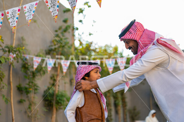 مفهوم الأناقة والاهتمام بالمظهر الخارجي، أجواء عائلية مليئة بالحب, الاحتفال بالمناسبات السعيدة في الهواء الطلق, ارتداء الأزياء التقليدية في المناسبات والأعياد الإسلاميه, صورة مقربة من الجانب لرجل عربي سعودي خليجي يرتدي الزي التقليدي يقوم بتعديل شماغ ابنه
