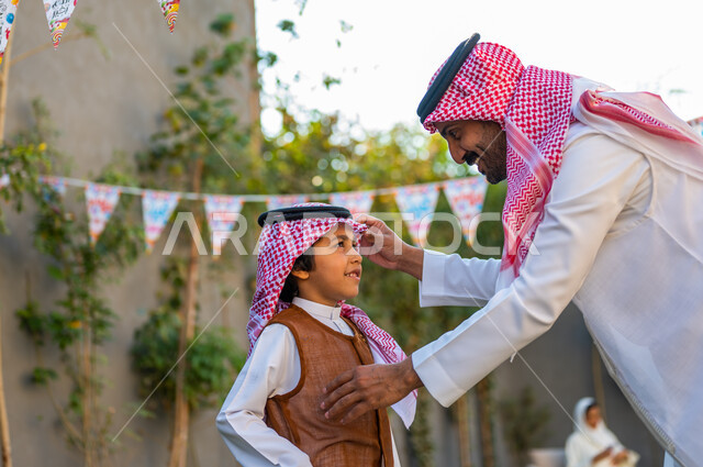 أجواء عائلية مليئة بالحب, الاحتفال بالمناسبات السعيدة في الهواء الطلق, صورة مقربة من الجانب لرجل عربي سعودي خليجي يرتدي الزي التقليدي يقوم بتعديل شماغ ابنه,  مفهوم الأناقة والاهتمام بالمظهر الخارجي، ارتداء الأزياء التقليدية في المناسبات والأعياد الإسلاميه