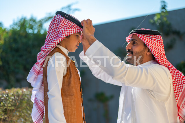  ارتداء الأزياء التقليدية في المناسبات والأعياد الإسلاميه، مفهوم الأناقة والاهتمام بالمظهر الخارجي, صورة مقربة من الجانب لرجل عربي سعودي خليجي يرتدي الزي التقليدي يقوم بتعديل شماغ ابنه, الاحتفال بالمناسبات السعيدة في الهواء الطلق, أجواء عائلية مليئة بالحب 
