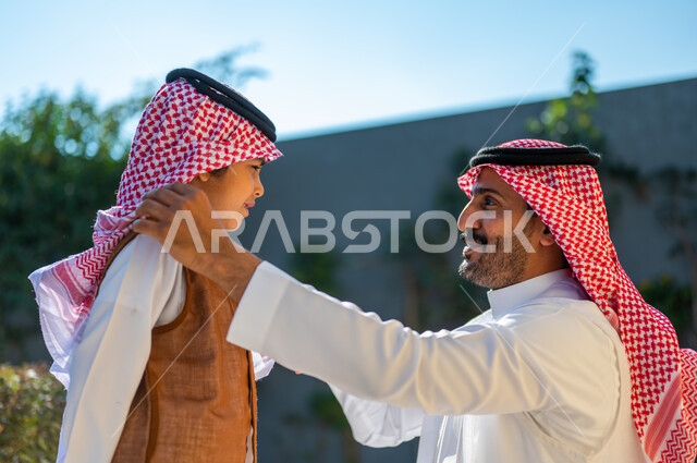 Celebrating happy occasions outdoors, wearing traditional costumes on Islamic occasions and holidays, a close-up photo from the side of a Saudi Gulf Arab man wearing traditional clothing adjusting his son’s shemagh, a family atmosphere full of love, the concept of elegance and attention to external appearance