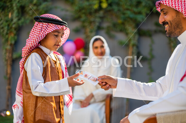طقوس العادات والتقاليد العربية في المناسبات والأعياد الاسلامية، قضاء وقت ممتع في أيام العيد في السعوديه، صورة من الجانب لفتى عربي سعودي خليجي يرتدي الملابس الجديدة ويأخذ العيدية من والده، إدخال الفرحة والبهجة لقلوب الأولاد
