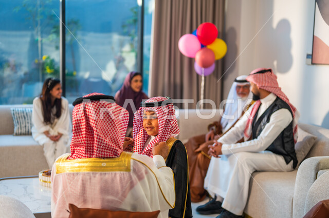 قضاء اوقات ممتعة مع الابناء، تعابير السعادة والسرور، صورة مقربة من الخلف لرجل يرتدي الزي التقليدي، أسرة عربيه خليجية سعودية تجلس في غرفة المعيشة تقوم بالاحتفال بالعيد