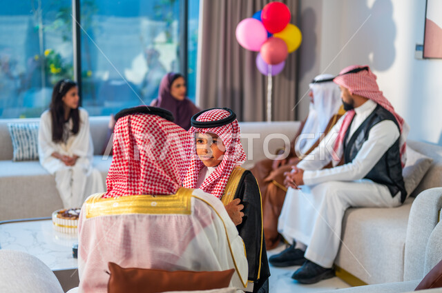 تعابير السعادة والسرور، صورة مقربة من الخلف لرجل يرتدي الزي التقليدي، أسرة عربيه خليجية سعودية تجلس في غرفة المعيشة تقوم بالاحتفال بالعيد، قضاء اوقات ممتعة مع الابناء