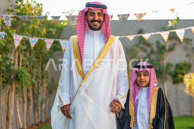 Wearing traditional costumes on Islamic occasions, the concept of paying attention to external appearance and elegance, decorations for happy holidays, standing cheerfully and happily in the garden of the house, a smiling Saudi Gulf Arab man wearing a shemagh and white abaya, holding his son’s hand happily