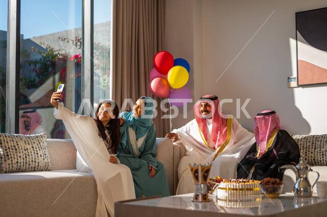 Eid Mubarak balloons and decorations, taking a selfie using a mobile phone, using advanced modern technologies, family gatherings and visits in the living room, the concept of family cohesion, gestures of cheerfulness, joy and happiness, a Saudi Arab family celebrating the advent of the Happy Eid