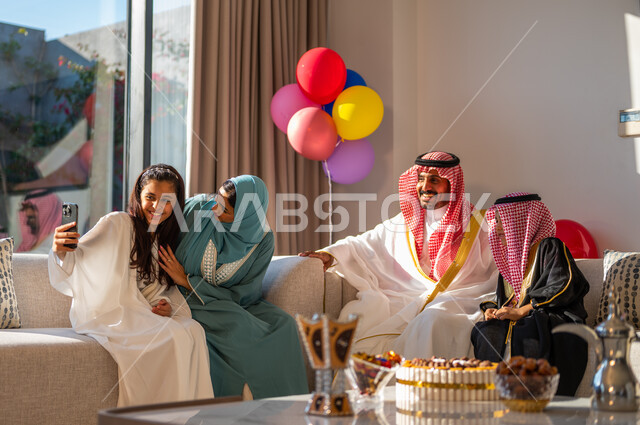 Eid Mubarak balloons and decorations, taking a selfie using a mobile phone, using advanced modern technologies, family gatherings and visits in the living room, the concept of family cohesion, gestures of cheerfulness, joy and happiness, a Saudi Arab family celebrating the advent of the Happy Eid