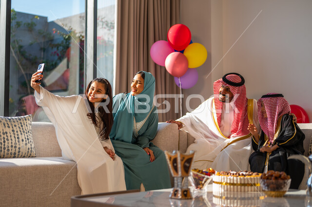 Eid Mubarak balloons and decorations, taking a selfie using a mobile phone, using advanced modern technologies, family gatherings and visits in the living room, the concept of family cohesion, gestures of cheerfulness, joy and happiness, a Saudi Arab family celebrating the advent of the Happy Eid