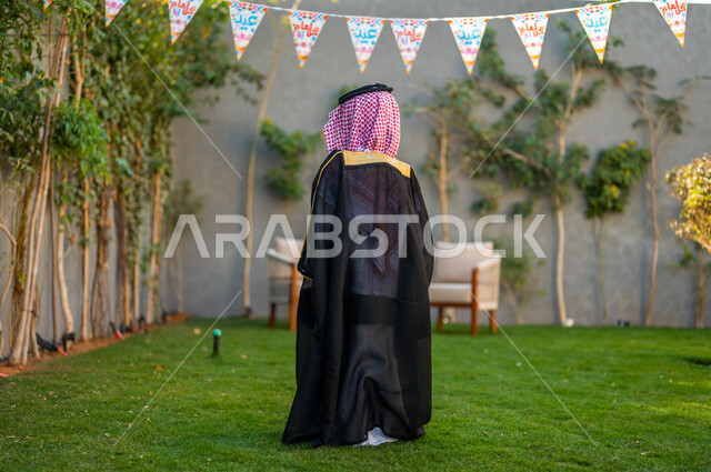  الاحتفال بالهواء الطلق, الاهتمام بالمظهر الخارجي للأطفال, صورة من الخلف لفتى عربي خليجي سعودي يرتدي البشت الأسود والعقال يستمتع في حديقة المنزل, زينة الأعياد والمناسبات الاسلامية, فرحة وبهجة الأولاد بالعيد المبارك