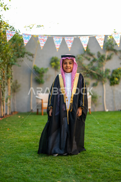 الاهتمام بالمظهر الخارجي للأطفال, فتى عربي خليجي سعودي مبتسم يرتدي البشت الأسود والعقال ينظر إلى الكاميرا ويستمتع في حديقة المنزل, زينة الأعياد والمناسبات الاسلامية, فرحة وبهجة الأولاد بالعيد المبارك, الاحتفال بالهواء الطلق