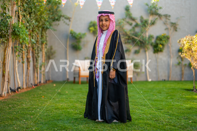 الاحتفال بالهواء الطلق,  فتى عربي خليجي سعودي يرتدي البشت الأسود والعقال ينظر إلى الكاميرا ويستمتع في حديقة المنزل, زينة الأعياد والمناسبات الاسلامية, الاهتمام بالمظهر الخارجي للأطفال, فرحة وبهجة الأولاد بالعيد المبارك